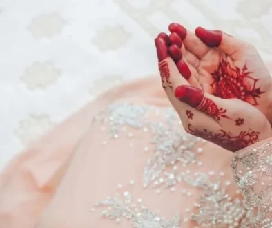 Close-up of a Muslim bride in an elegant light peach dress with henna-adorned hands raised in du'a prayer on a white patterned carpet.