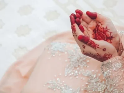 Close-up of a Muslim bride in an elegant light peach dress with henna-adorned hands raised in du'a prayer on a white patterned carpet.