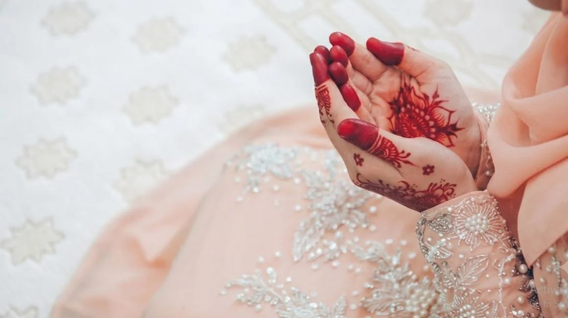 Close-up of a Muslim bride in an elegant light peach dress with henna-adorned hands raised in du'a prayer on a white patterned carpet.